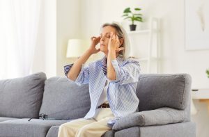 Sick mature woman sitting on sofa, suffering from headache or hypertension, rubbing her temples. Tired pensioner with high blood pressure feeling exhausted and unwell. Cardiovascular diseases concept.